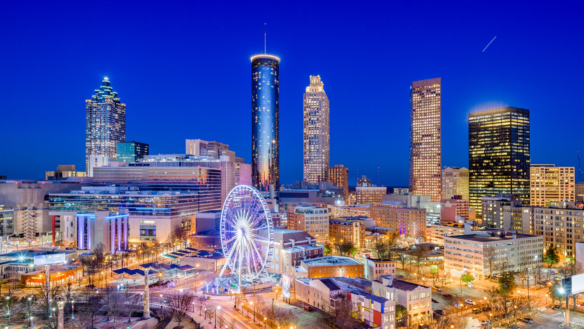 A vibrant evening cityscape of downtown Atlanta, Georgia, featuring illuminated skyscrapers, the glowing SkyView Ferris wheel, and bustling streets below, all under a deep blue twilight sky.