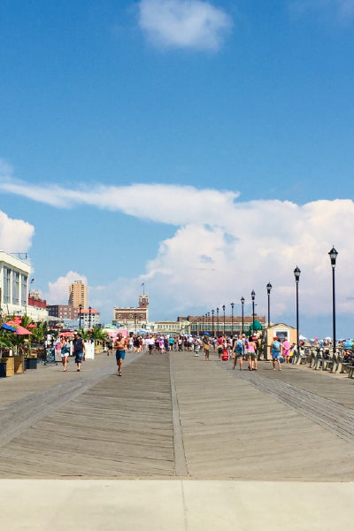 A sunny day at a lively boardwalk crowded with people walking and enjoying the outdoors, lined with shops and street lamps, under a bright blue sky with scattered clouds.