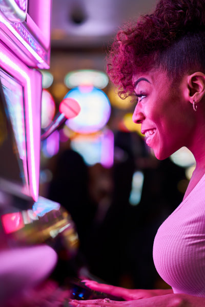 A close-up profile of a smiling young woman with curly hair playing a brightly lit slot machine in a casino, surrounded by colorful, blurred lights in the background.