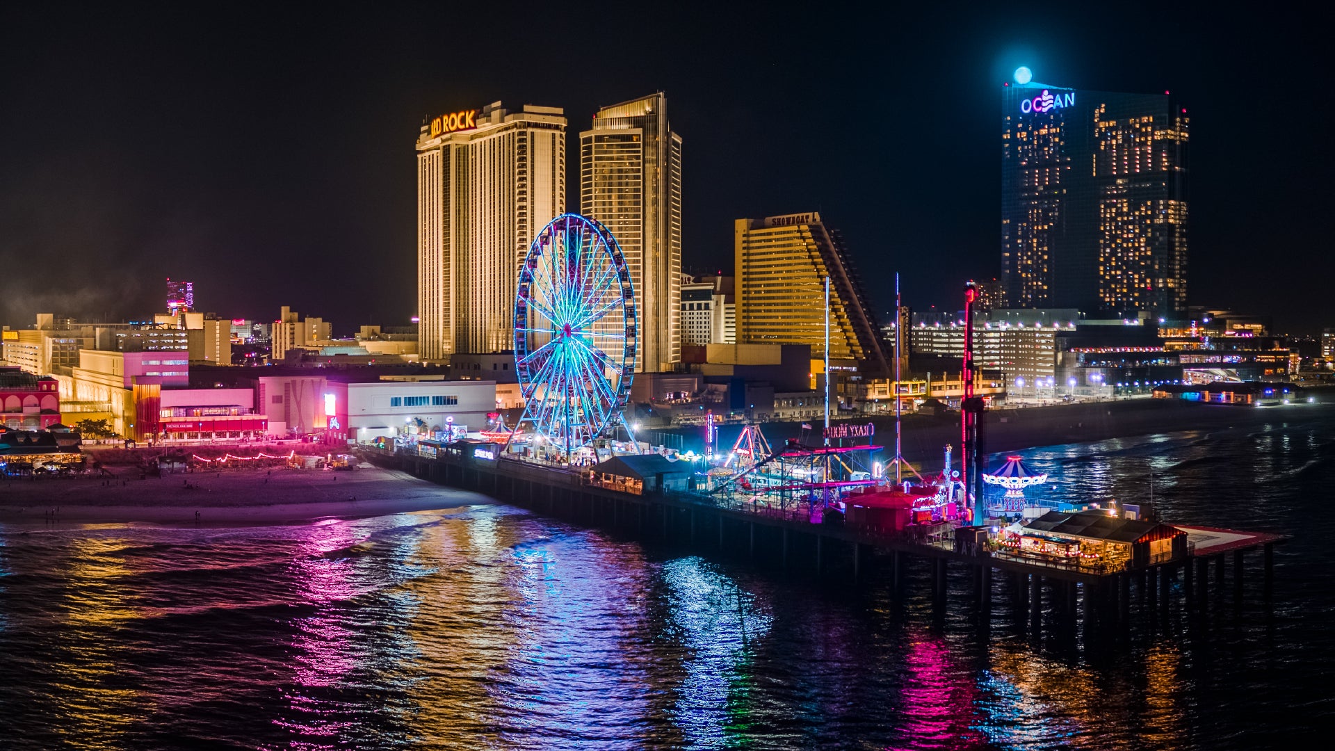 A vibrant nighttime view of the Atlantic City boardwalk and pier, featuring a brightly lit Ferris wheel, colorful amusement rides, and the illuminated facades of prominent casinos and hotels, with reflections shimmering on the ocean water.