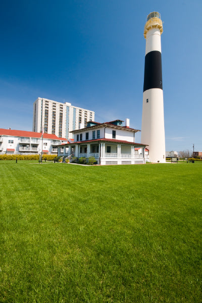 A tall lighthouse with a black and white striped pattern stands next to a white building with a red roof, surrounded by a large, well-maintained green lawn under a clear blue sky. In the background, there is a multi-story building.