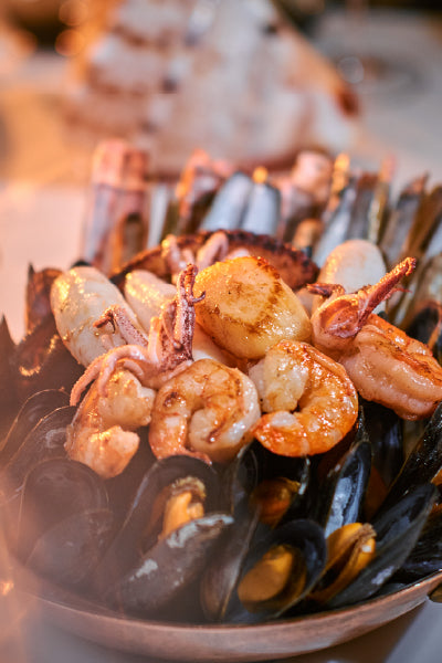 A close-up of a seafood platter featuring grilled shrimp, scallops, and baby squid arranged on a bed of mussels, with steam rising, suggesting the dish is freshly cooked and served hot.