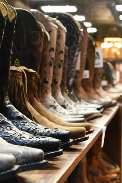 Row of various cowboy boots displayed on a wooden shelf in a store, showcasing different styles, colors, and designs.