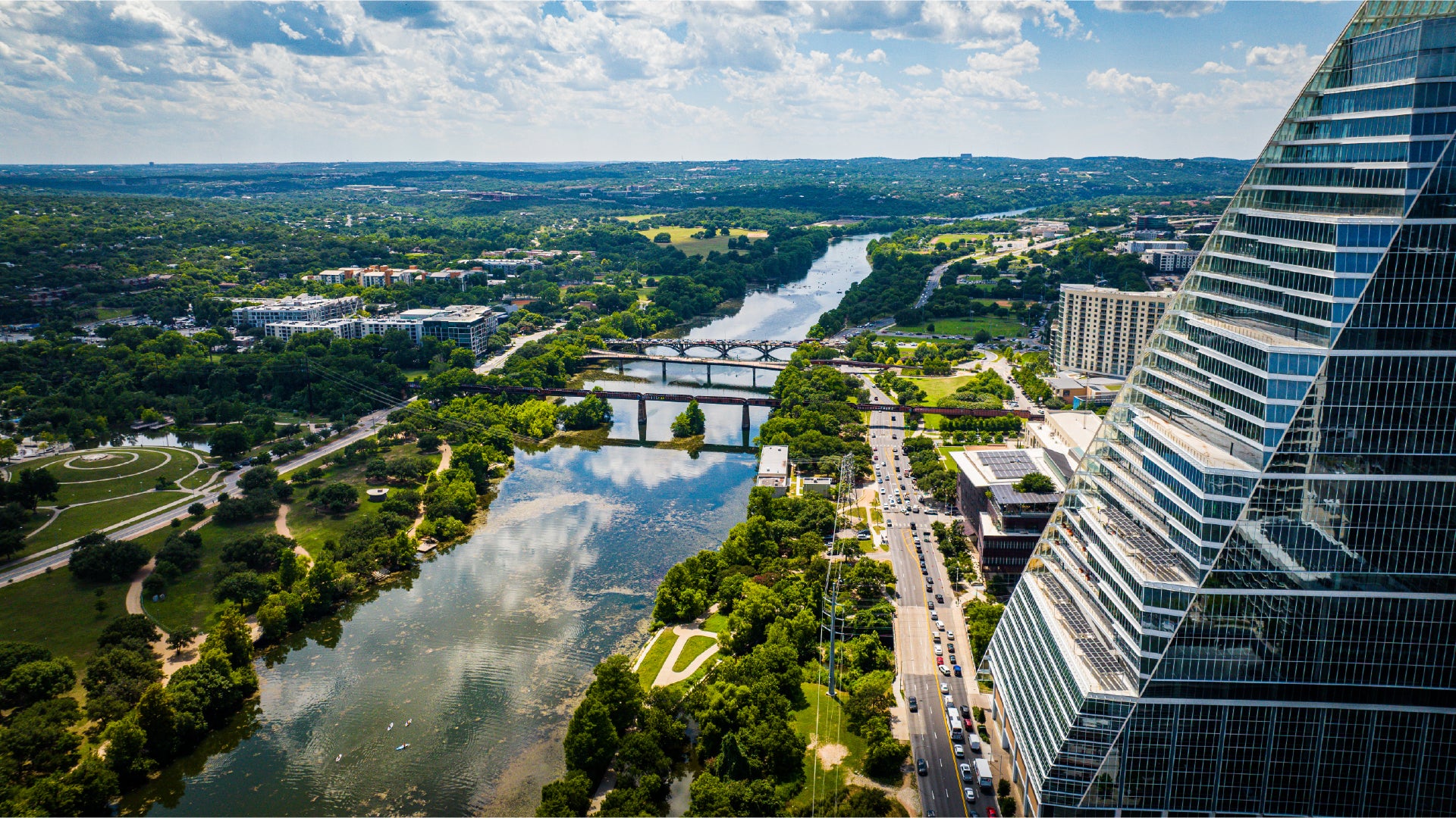 Aerial view of a cityscape featuring a large river running through a green park area with multiple bridges crossing the river, and a modern glass skyscraper on the right side reflecting the sky. The scene is set on a sunny day with a partly cloudy blue sky.