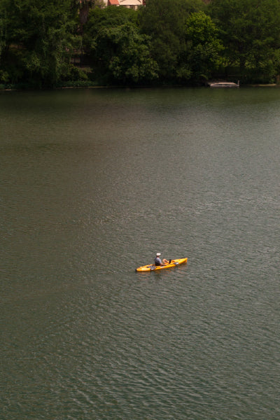 Person paddling a yellow kayak on a calm river, surrounded by lush green trees along the riverbank.
