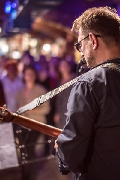 Man playing guitar on stage at a live music event, with a blurred audience watching in the background under soft, colorful lighting.