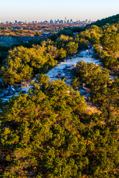 Scenic aerial view of a nature trail winding through dense green trees with several people walking and enjoying the outdoors, and a distant city skyline visible on the horizon under a clear blue sky.