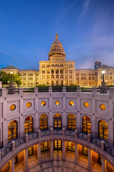 Evening view of the Texas State Capitol building illuminated with warm lights against a clear blue sky, seen from inside a circular, colonnaded courtyard with arched doorways and round windows.