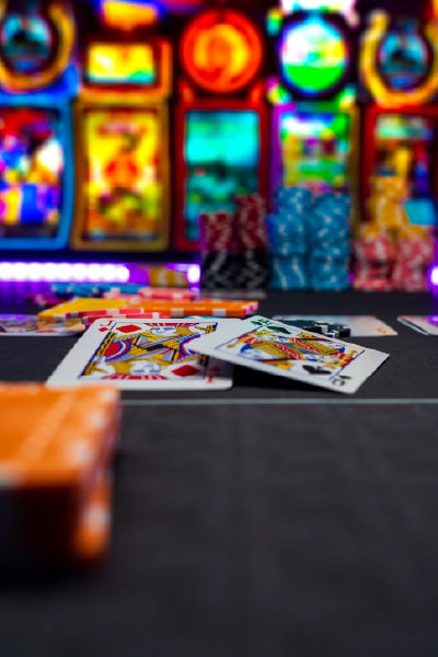 A close-up view of a poker table with playing cards and colorful poker chips scattered on it. In the background, brightly lit and blurred slot machines create a vibrant casino atmosphere.