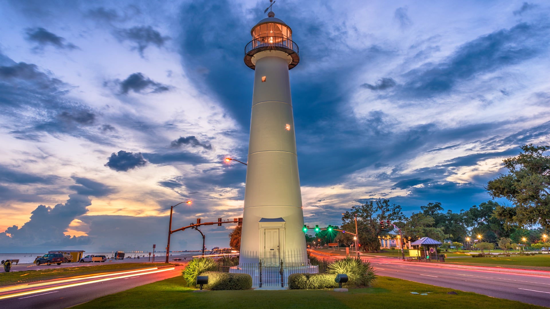 A tall white lighthouse stands illuminated at dusk with its light glowing near the top. The sky is filled with dramatic clouds and colors of sunset, while car light trails blur along the road on either side of the lighthouse. Trees and streetlights line the area, creating a serene coastal scene.