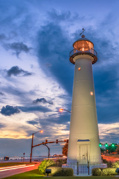 A tall white lighthouse stands illuminated at dusk with its light glowing near the top. The sky is filled with dramatic clouds and colors of sunset, while car light trails blur along the road on either side of the lighthouse. Trees and streetlights line the area, creating a serene coastal scene.