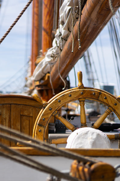 Close-up of a wooden ship's steering wheel on the deck of a sailboat, with ropes, masts, and sails visible in the background under a clear sky.