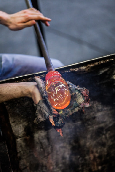 A person shaping molten glass on a blowpipe using a protective pad, with the glowing hot glass being carefully manipulated in a glassblowing workshop.