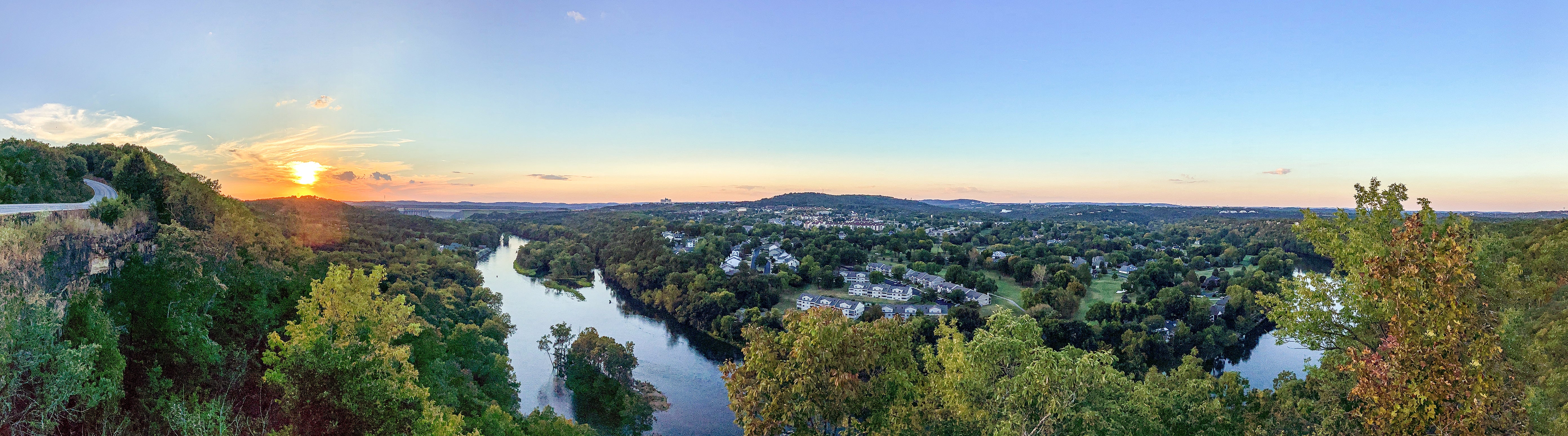 A panoramic view of a lush, green valley with a winding river running through it, surrounded by dense trees and suburban homes. The sun is setting on the left side of the image, casting a golden glow across the sky and reflecting on the water. A road curves along the edge of a cliff on the far left, while the right side shows expansive neighborhoods nestled in the forest. The sky transitions from warm orange near the horizon to clear blue above, indicating a peaceful, late summer or early autumn evening.