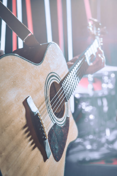 Close-up of an acoustic guitar being played, with bright stage lights and blurred drum set in the background.
