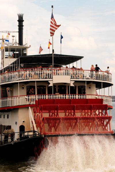 A large red and white paddlewheel riverboat cruising on the water with people standing on the upper deck, and an American flag flying at the top.