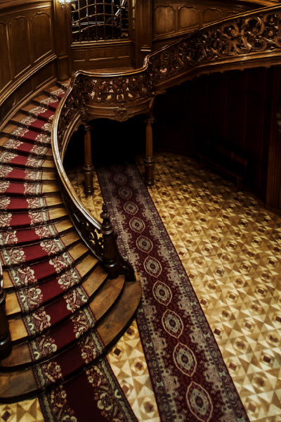 A grand, curved wooden staircase with an ornate carved banister and a red patterned carpet runner, leading down to a polished wooden floor with a matching decorative rug in an elegant interior setting.