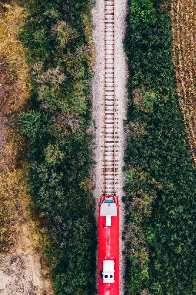 Aerial view of a red train traveling along a single railroad track surrounded by dense green vegetation and dry grass on either side.