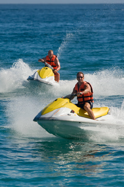 Two men wearing orange life jackets riding yellow and white jet skis on the ocean, creating splashes of water as they speed across the surface.