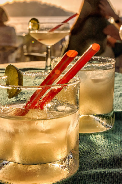 Two glasses of iced cocktails with salted rims, garnished with lime wedges and red straws, placed on a table with a blurred sunset and people in the background.