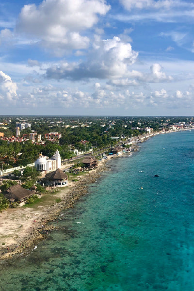 Aerial view of a coastal town with clear turquoise waters along a rocky shoreline, small buildings, a white church with a dome, and lush green vegetation under a partly cloudy blue sky.
