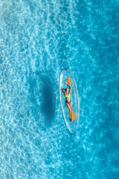 A woman wearing a green bikini relaxing in a clear, transparent kayak with an orange kayak oar floating on bright blue, crystal-clear water, viewed from above.