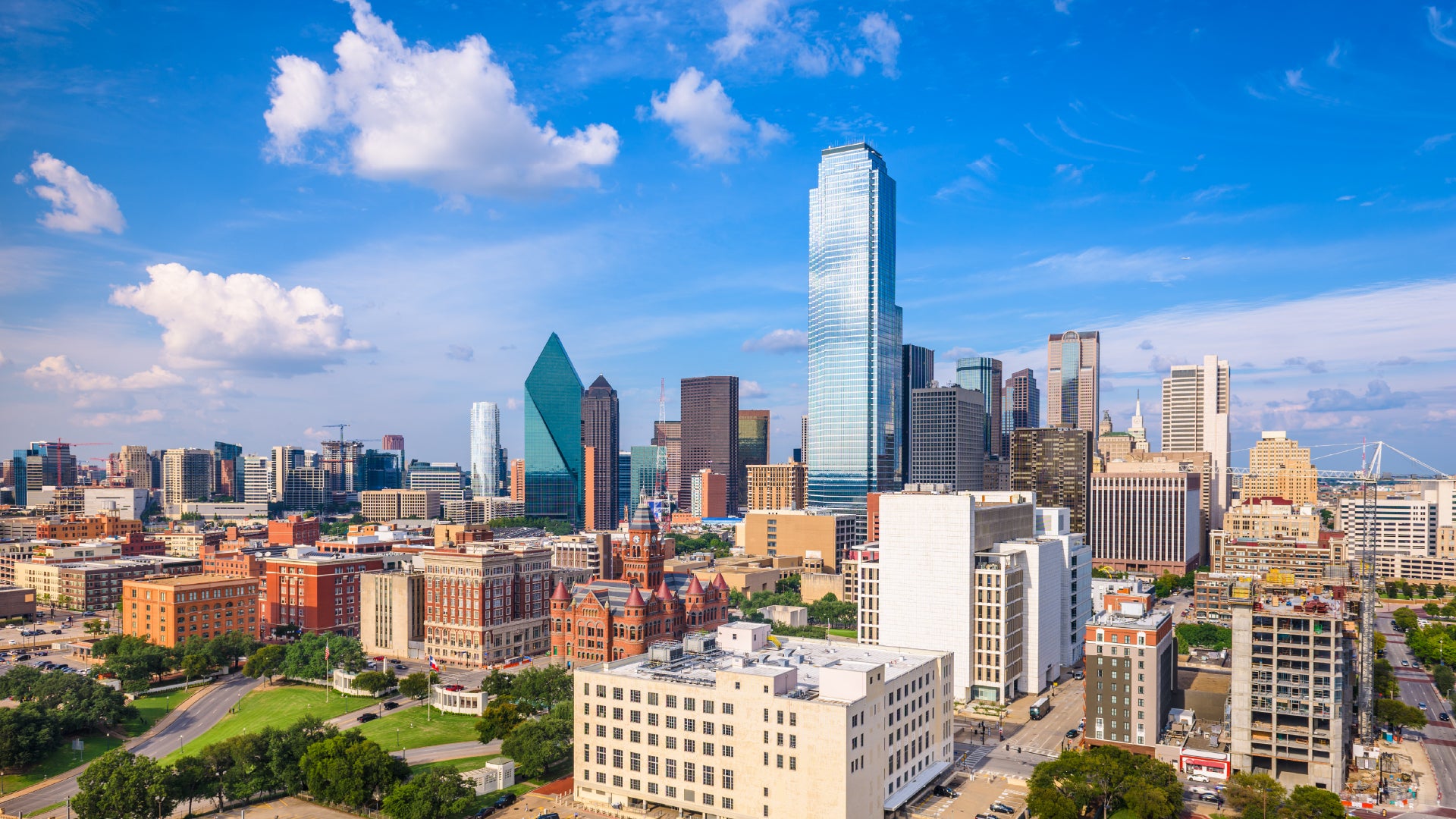 A vibrant view of downtown Dallas, Texas, showcasing modern skyscrapers, historic buildings, and green spaces under a bright blue sky. Iconic glass high-rises, including Fountain Place, contrast with the red-brick Old Red Museum, highlighting the city's blend of history and growth.