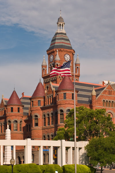 Historic red-brick courthouse building with a clock tower and conical turrets, featuring an American flag waving in the foreground under a partly cloudy sky.