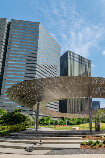 Modern urban park featuring a large, circular metallic canopy structure with perforated patterns, set against the backdrop of tall glass and steel office buildings under a clear blue sky.