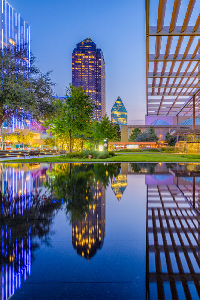 Evening cityscape featuring modern high-rise buildings with colorful lights reflecting in a still water feature, surrounded by trees and contemporary architectural structures in an urban park setting.