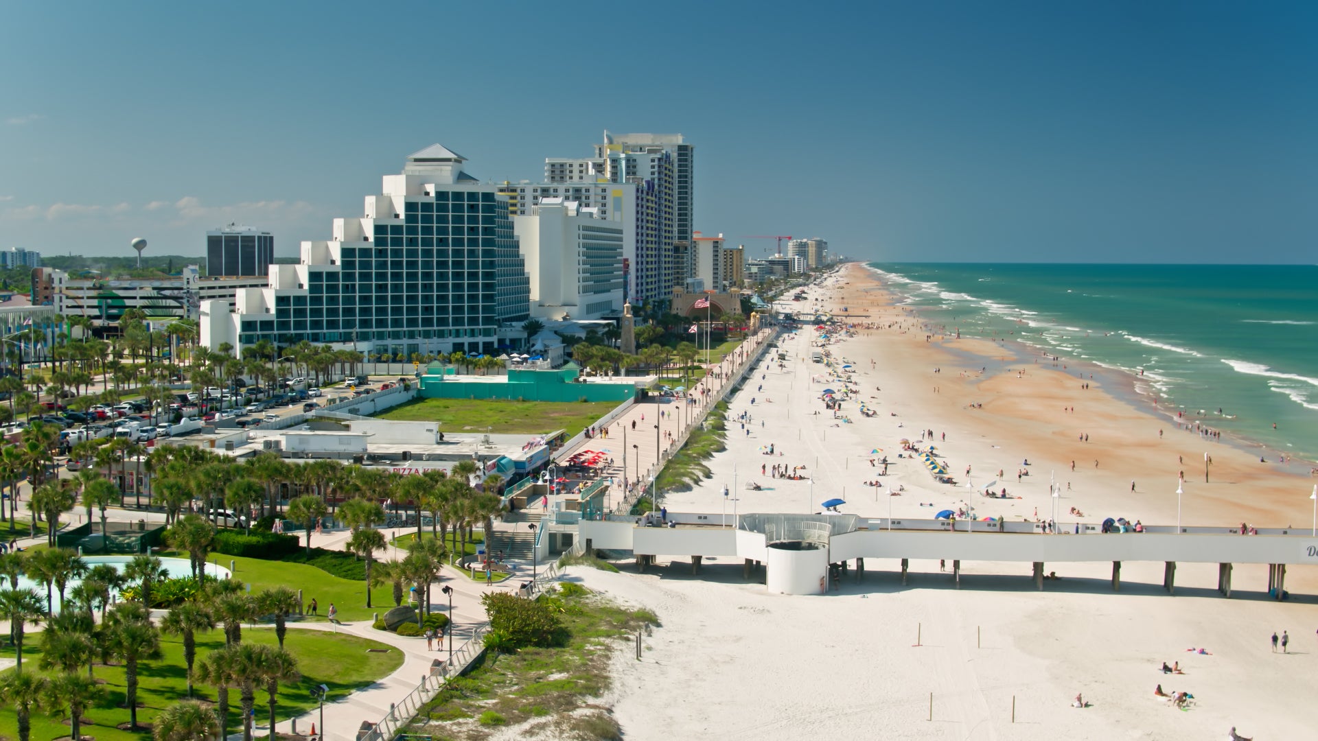 A scenic daytime view of Daytona Beach, Florida, showcasing its long sandy shoreline and turquoise ocean waves. The beachfront is lined with high-rise hotels and resorts, including a distinctively stepped pyramid-style building. The boardwalk and pier extend into the beach area, with people strolling, sunbathing, and enjoying the warm weather. Green spaces with palm trees and walkways add to the coastal charm of this popular vacation destination.