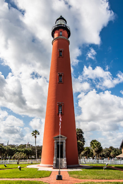 Tall, red brick lighthouse with small windows and a black lantern room at the top, standing against a partly cloudy blue sky, surrounded by palm trees and a white picket fence.