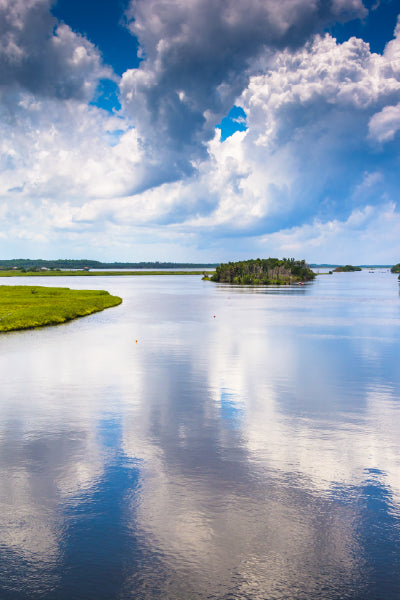 Calm river or lake with reflective water, small green islands, and lush grass along the shore under a dramatic sky filled with large, fluffy clouds.