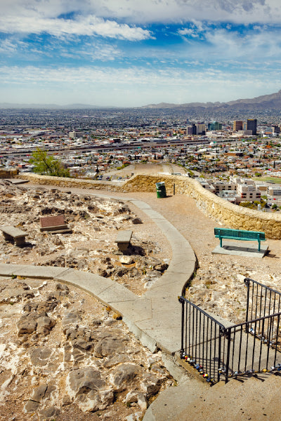 View from a rocky hilltop park with paved walkways, benches, and railings overlooking a sprawling cityscape with numerous buildings and mountains in the distance under a partly cloudy sky.