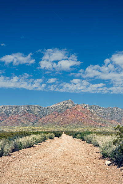 Dirt road stretching toward colorful red and gray mountains under a bright blue sky dotted with white clouds, surrounded by desert shrubs and grass.