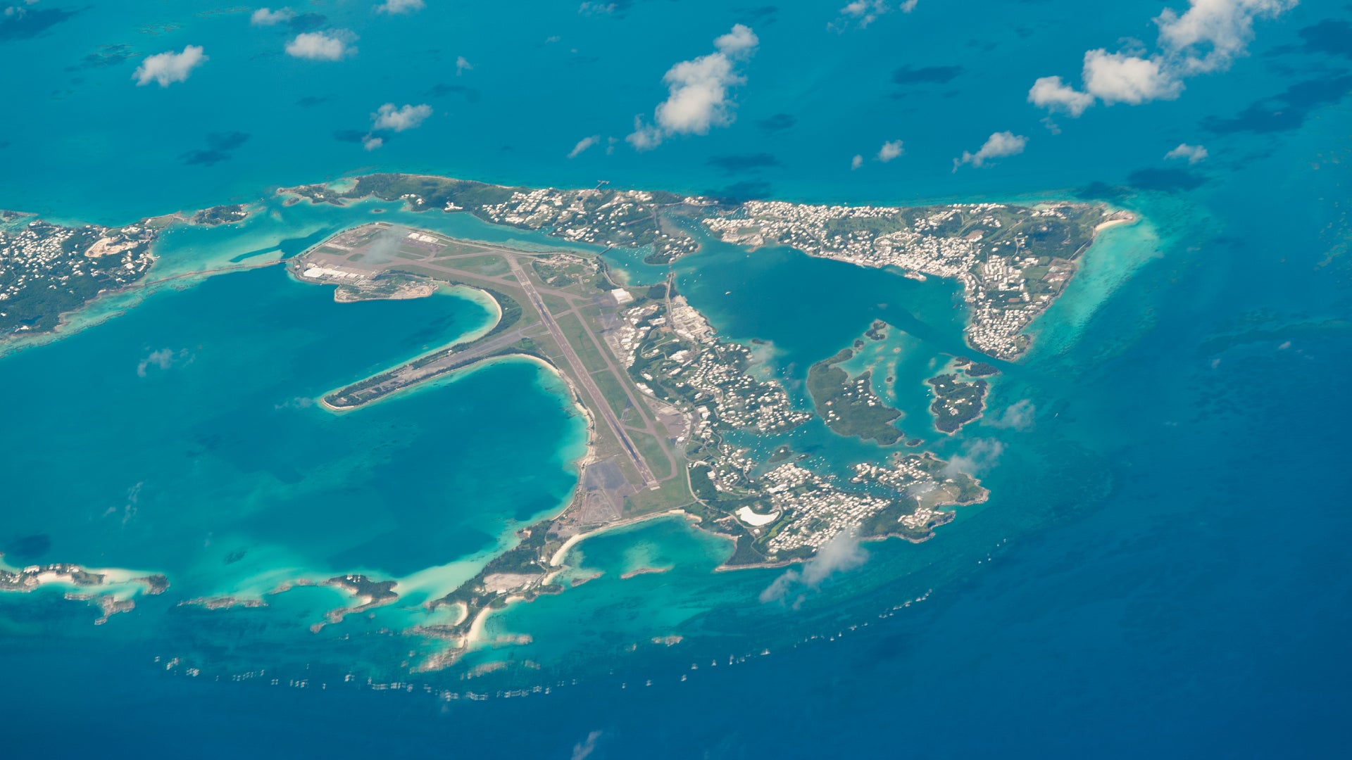 Aerial view of Freeport, Grand Bahama. The image prominently features L.F. Wade International Airport, visible with its long runway extending into the ocean on the left-central part of the image. Surrounding the airport are turquoise waters and coral reefs. The islands' densely populated areas and characteristic white-roofed buildings are also visible.