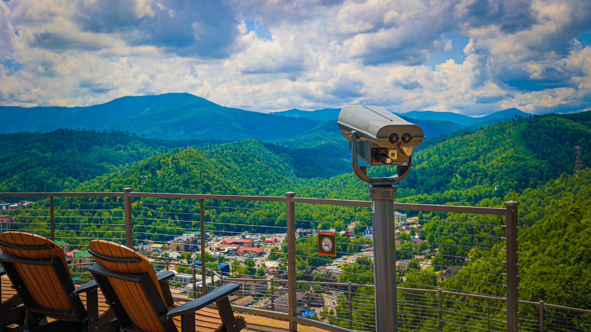 A breathtaking scenic view from a mountaintop observation deck overlooking a lush green valley and distant blue mountains. A coin-operated binocular viewer stands at the edge of the deck, inviting visitors to take a closer look at the picturesque landscape. Wooden lounge chairs face the view, offering a peaceful spot to relax. Below, a small town with red-roofed buildings nestles among the rolling hills. The sky is filled with fluffy white clouds, casting soft shadows over the terrain, enhancing the serene