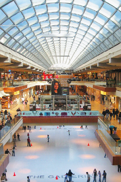 Indoor view of a shopping mall with a large arched glass ceiling, multiple levels of stores, and an ice skating rink on the lower level. Shoppers walk along the upper levels, while people skate on the rink below. A large advertisement with a woman's face hangs prominently in the center above the rink.