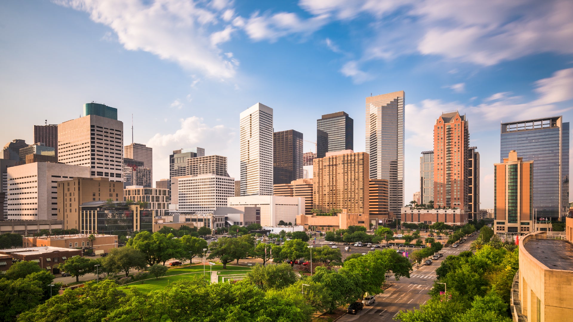 A bright and clear view of the Houston, Texas skyline, showcasing a mix of modern skyscrapers and contemporary buildings. The foreground features lush green trees and a small urban park, while the background consists of a blue sky with wispy clouds. The city's downtown district is bustling with commercial and residential structures, reflecting Houston's dynamic and growing metropolitan landscape.