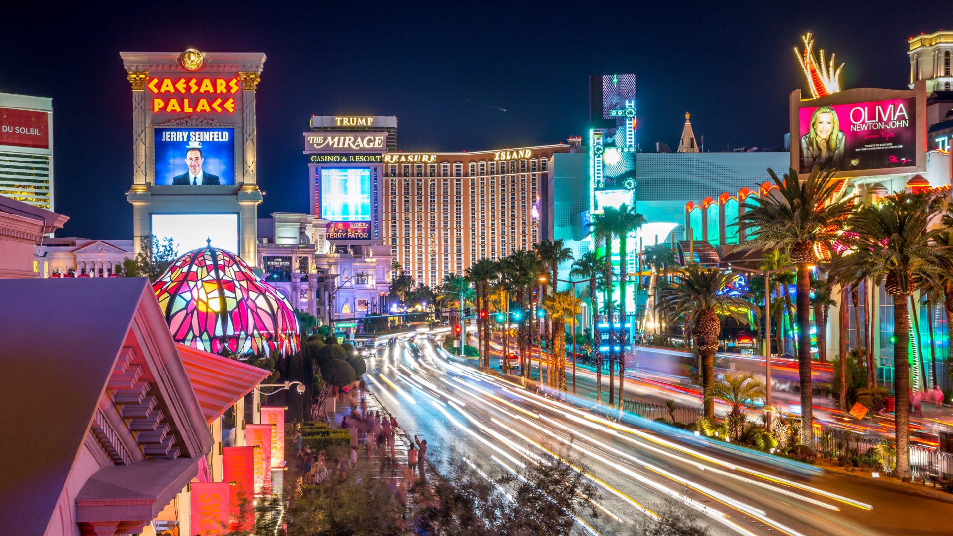 A vibrant nighttime view of the Las Vegas Strip, featuring bright neon lights, towering hotels, and bustling traffic. The illuminated Caesars Palace sign and The Mirage hotel stand prominently, alongside various colorful billboards advertising entertainers and shows. Palm trees line the busy street, where streaks of light from moving vehicles create a dynamic energy. Crowds of people can be seen walking along the sidewalks, soaking in the lively atmosphere of the city.