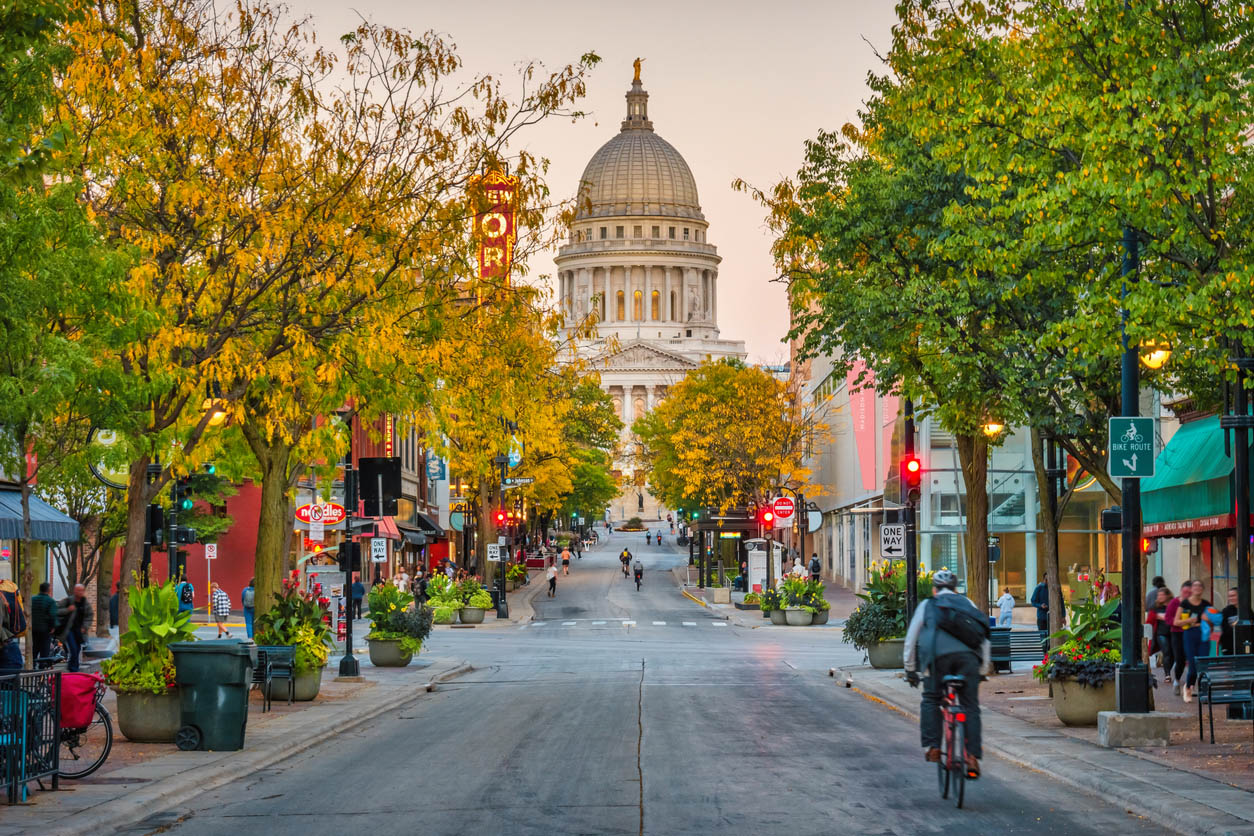 A vibrant city street scene leading to a grand domed capitol building in the background, framed by trees with golden autumn leaves. The street is lined with shops, cafes, and colorful signs, with pedestrians walking along the sidewalks and a cyclist riding towards the camera. The atmosphere is lively, with warm lighting from street lamps and storefronts, creating a welcoming urban setting. The capitol building stands as a striking focal point, adding a sense of history and grandeur to the scene.