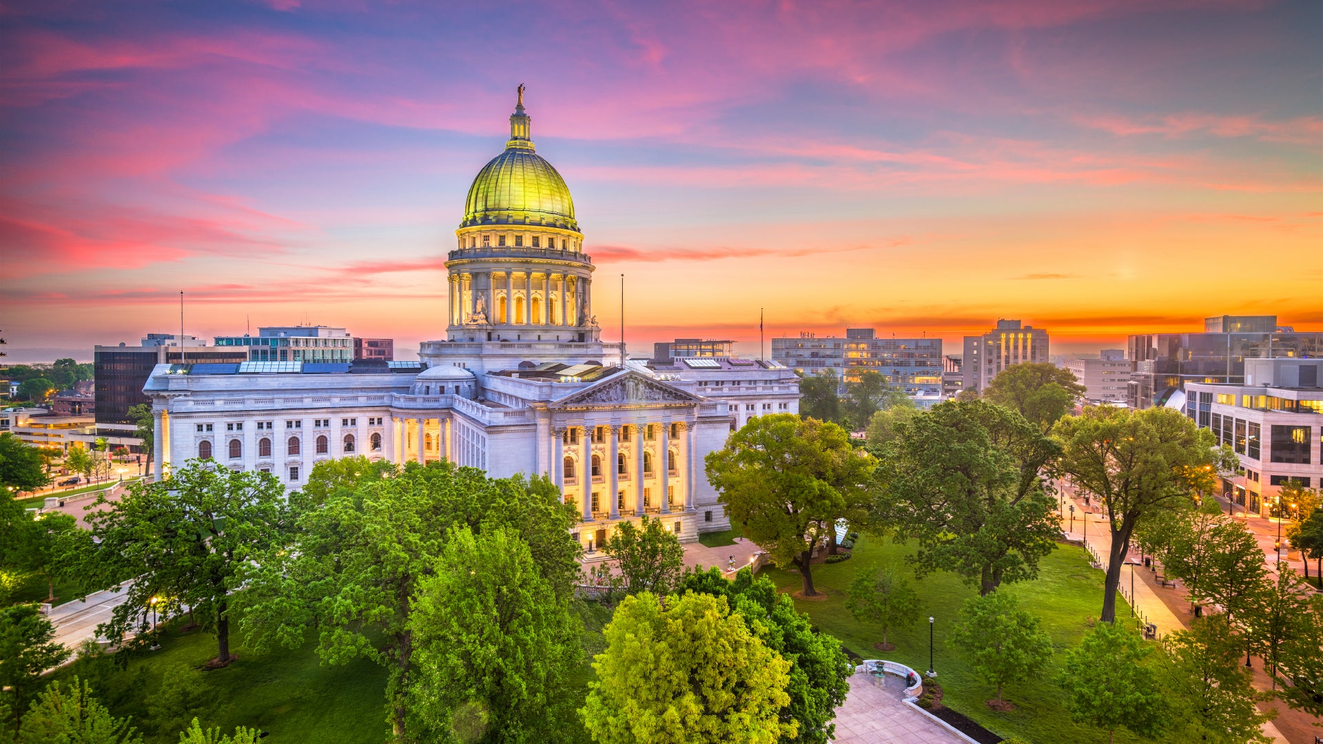 A grand capitol building with a golden-domed roof stands illuminated against a stunning sunset sky with shades of pink, orange, and purple. The neoclassical architecture features towering columns and intricate details, surrounded by lush green trees and a well-manicured park. The cityscape in the background includes modern buildings, creating a beautiful contrast between history and contemporary urban life.