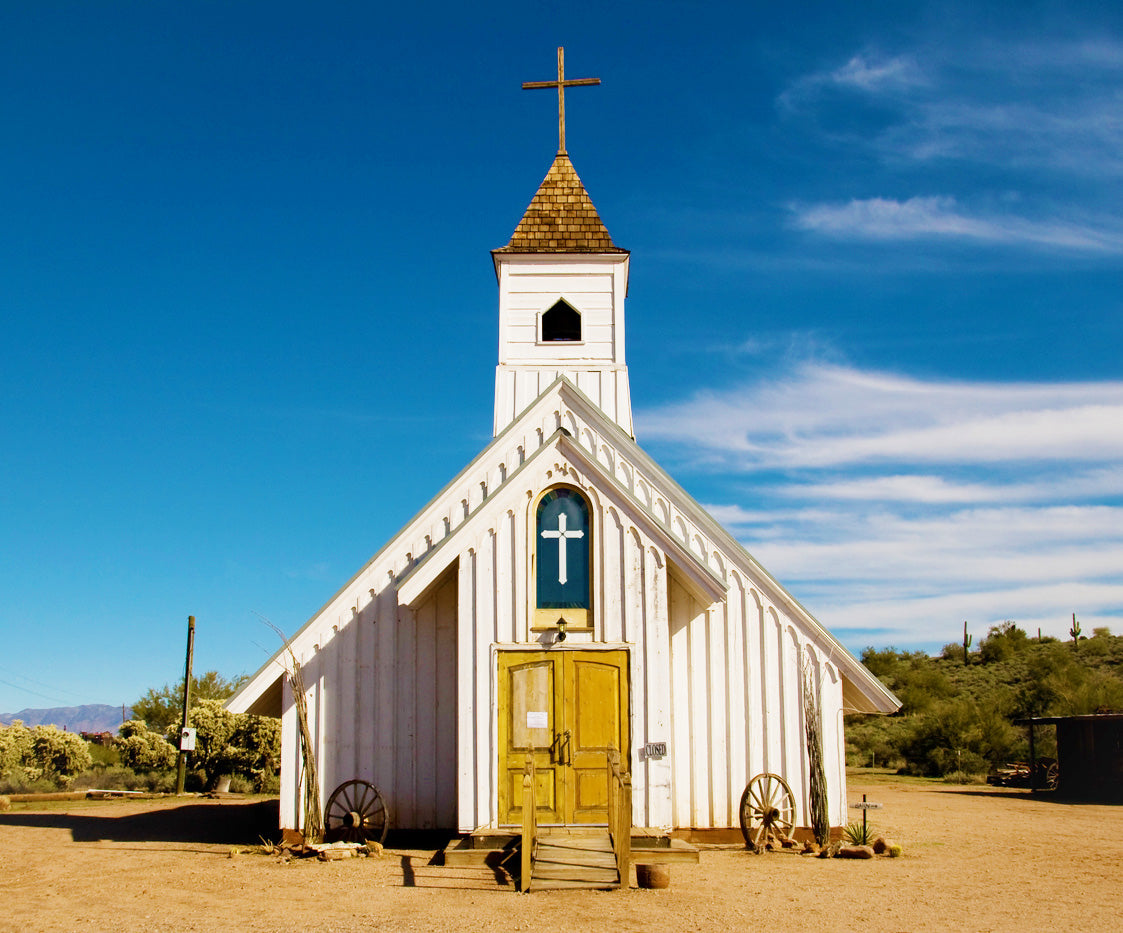 A small, rustic white wooden church with a bell tower and cross, set in a desert landscape under a clear blue sky. The weathered wooden doors and arched window with a cross add to its historic charm, with wagon wheels and sparse desert vegetation surrounding the building.