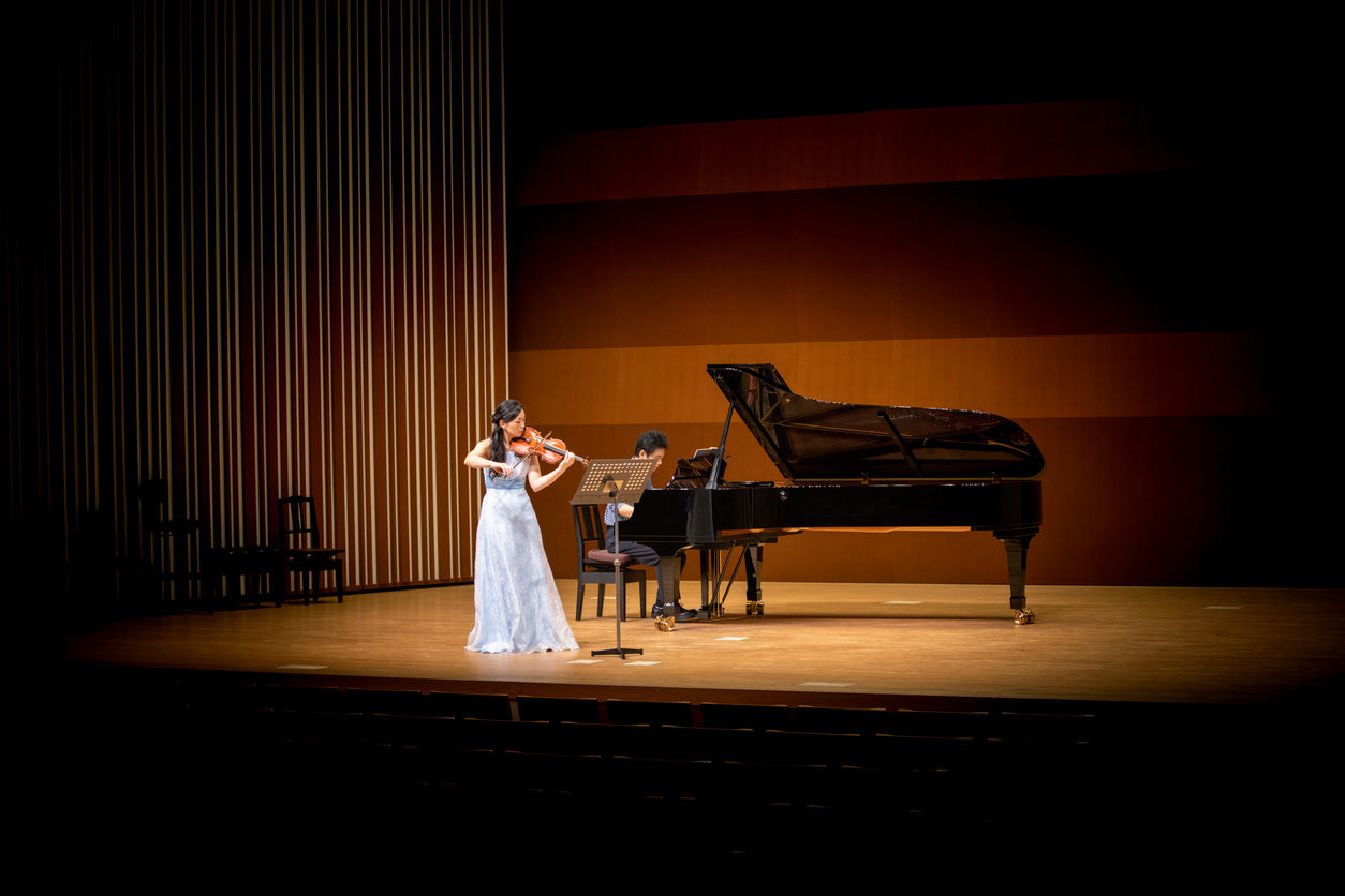 A violinist in an elegant white gown performs on stage alongside a pianist dressed in black, who is seated at a grand piano. The stage is illuminated with warm lighting, and the background features a wooden panel design with vertical and horizontal elements. The performers are focused on their music, with a music stand positioned between them. The audience seating is visible in the foreground, though darkened.