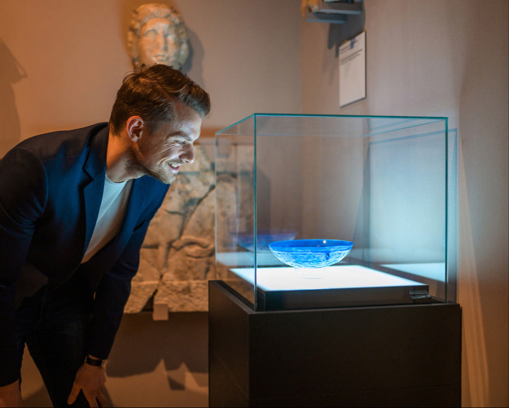 A man with light skin, short brown hair, and a beard, wearing a navy blue blazer and white shirt, is leaning forward with a smile, closely examining a vibrant blue glass bowl displayed in a museum exhibit. The bowl is placed on a white-lit pedestal inside a transparent glass case. The background features ancient stone artifacts and sculptures on the wall, creating a contrast between the modern display and historical surroundings.