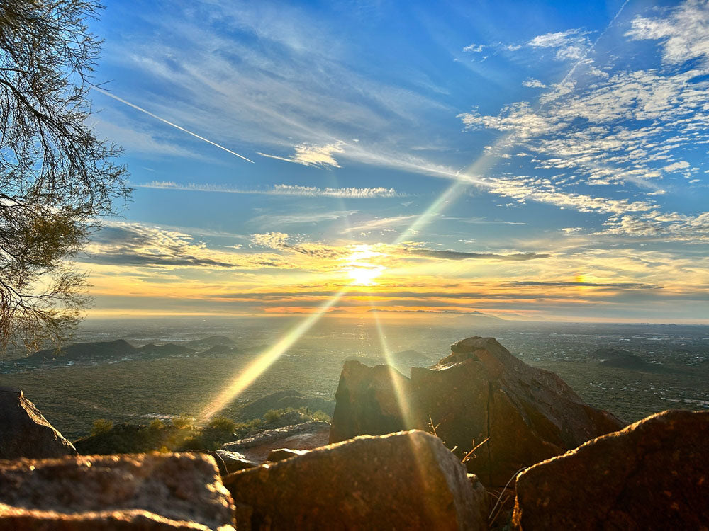 A breathtaking sunset view from a rocky mountain overlook, with the sun casting golden light across the horizon. The sky is a mix of soft blues and scattered clouds, with sun rays forming a striking X-shaped flare. Rolling hills and a distant cityscape stretch out below, while rugged rocks and a tree frame the scene, adding depth and texture.