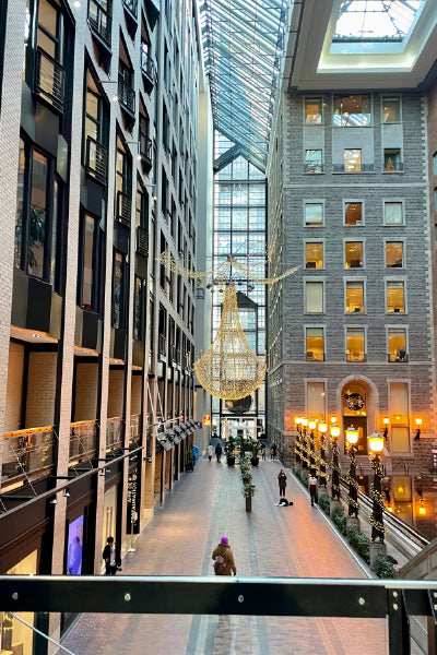 Interior view of a glass-roofed atrium in a city building, featuring a large, ornate chandelier suspended in the center. The space is flanked by tall buildings with windows, creating an alley-like corridor below. People walk along the brick-paved path lined with decorative street lamps and holiday garlands, suggesting a festive season.