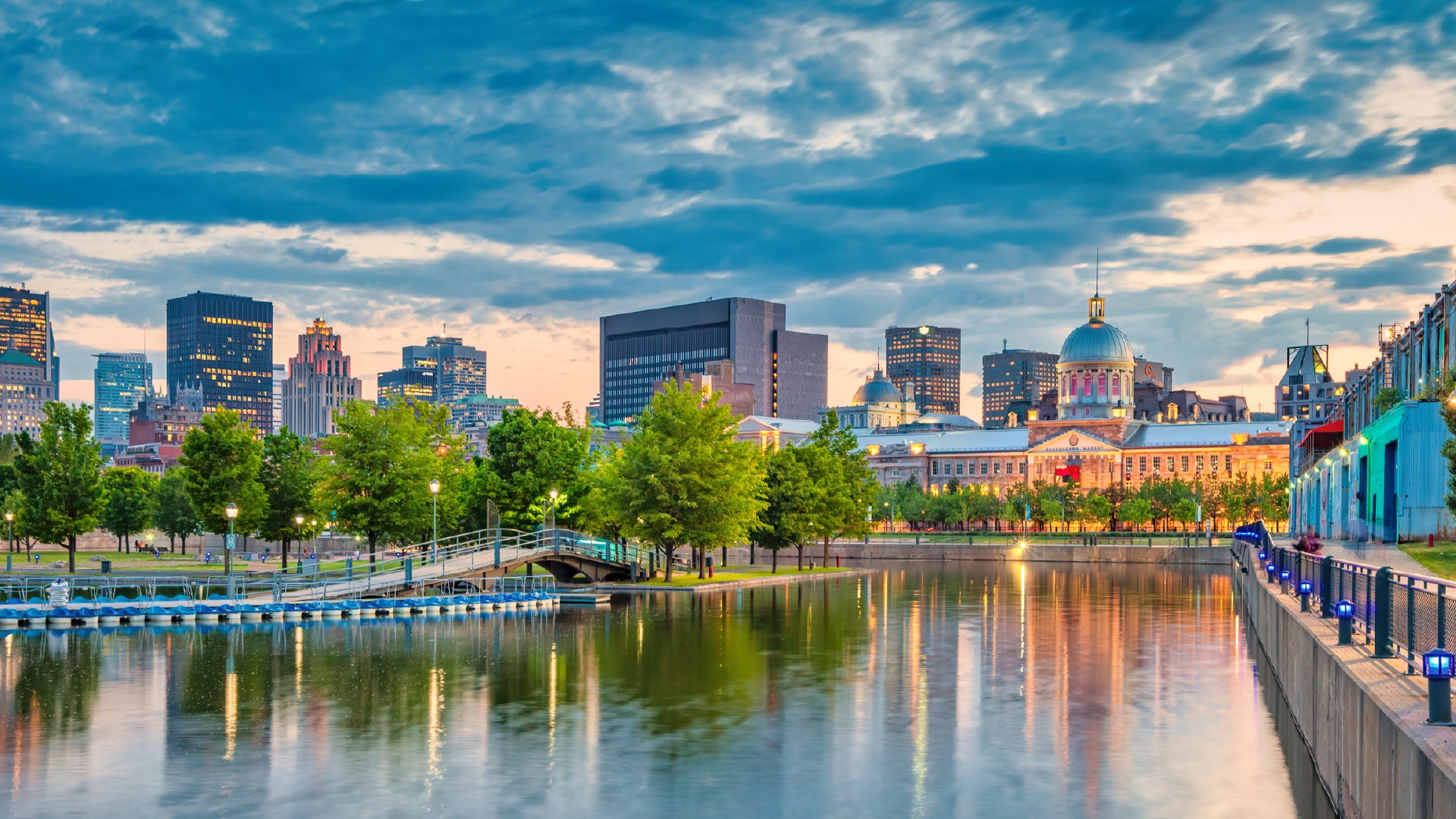 A stunning cityscape of Montreal, Canada, at dusk, featuring the historic Bonsecours Market with its illuminated dome and a blend of modern and classical architecture in the skyline. The foreground showcases a calm waterfront reflecting the city lights, with a pedestrian bridge and a tree-lined promenade adding charm to the scene. The sky is painted with hues of blue and orange, creating a picturesque urban setting.