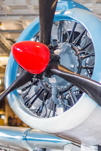Close-up view of a vintage aircraft's radial engine and propeller, featuring a shiny red spinner and black blades. The exposed mechanical components are surrounded by a light blue and white engine cowling, with the scene set indoors under bright lighting.