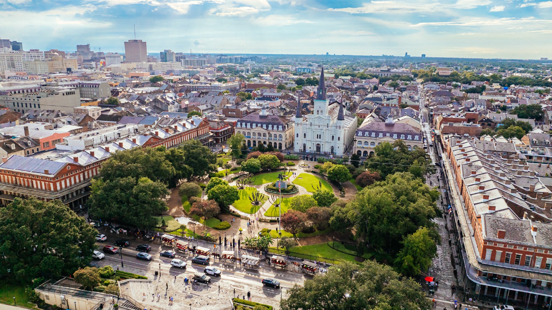 Aerial view of Jackson Square in New Orleans' French Quarter, featuring the St. Louis Cathedral, lush gardens, and an equestrian statue of Andrew Jackson. Historic buildings with colonial architecture surround the square, while horse-drawn carriages and cars line Decatur Street. The downtown skyline is visible in the background.